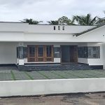 A new single-storey home, 3 BHK, is for sale in a gated villa complex in Thiruvalla, Kerala. The home has a white exterior, a spacious front yard with green pavers and gravel, a Sweetwater well and a low white boundary wall with a gate.