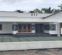 A new single-storey home, 3 BHK, is for sale in a gated villa complex in Thiruvalla, Kerala. The home has a white exterior, a spacious front yard with green pavers and gravel, a Sweetwater well and a low white boundary wall with a gate.