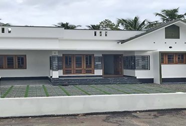 A new single-storey home, 3 BHK, is for sale in a gated villa complex in Thiruvalla, Kerala. The home has a white exterior, a spacious front yard with green pavers and gravel, a Sweetwater well and a low white boundary wall with a gate.