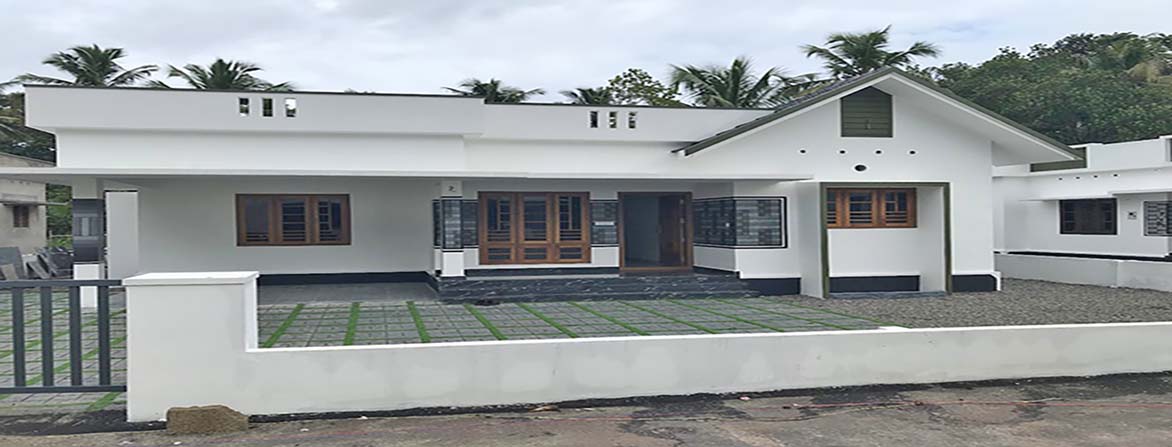 A new single-storey home, 3 BHK, is for sale in a gated villa complex in Thiruvalla, Kerala. The home has a white exterior, a spacious front yard with green pavers and gravel, a Sweetwater well and a low white boundary wall with a gate.