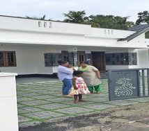 A newly built, modern single-storey home in Thiruvalla, Kerala, featuring a white exterior, a gated entrance with a grey decorative metal gate, and a front yard with green paver blocks. A tenant family of two women and a young girl are standing in the driveway. Their faces are blurred.