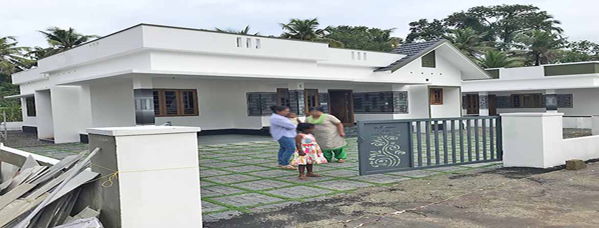 A newly built, modern single-storey home in Thiruvalla, Kerala, featuring a white exterior, a gated entrance with a grey decorative metal gate, and a front yard with green paver blocks. A tenant family of two women and a young girl are standing in the driveway. Their faces are blurred.