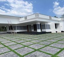 A newly constructed, modern single-storey home in Thiruvalla, Kerala, with a white exterior and a distinctive patterned front yard featuring grey square pavers with green grass in between. The house has a covered car porch on the left, an inviting entrance with dark steps, and a clean, contemporary design under a partly cloudy sky. A ladder is propped against the house near the porch.