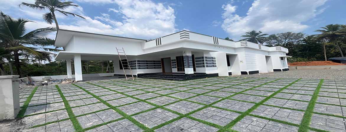 A newly constructed, modern single-storey home in Thiruvalla, Kerala, with a white exterior and a distinctive patterned front yard featuring grey square pavers with green grass in between. The house has a covered car porch on the left, an inviting entrance with dark steps, and a clean, contemporary design under a partly cloudy sky. A ladder is propped against the house near the porch.