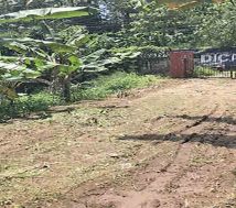 A view of the entrance to a commercial land for sale near Manjadi, Thiruvalla, with a metal gate flanked by two brick pillars and a banner for a medical college in the background.