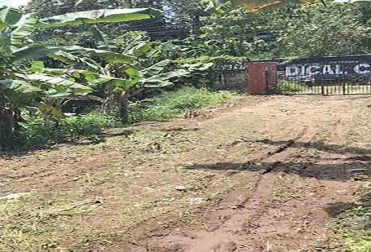 A view of the entrance to a commercial land for sale near Manjadi, Thiruvalla, with a metal gate flanked by two brick pillars and a banner for a medical college in the background.