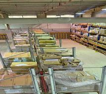 An overhead interior view of a vast industrial warehouse, showcasing its impressive 11-meter height. The concrete floor is clean, with long aisles separating rows of tall, heavy-duty metal racking systems filled with palletized goods wrapped in plastic. Bright overhead lighting illuminates the space, and exposed ductwork runs along the ceiling.