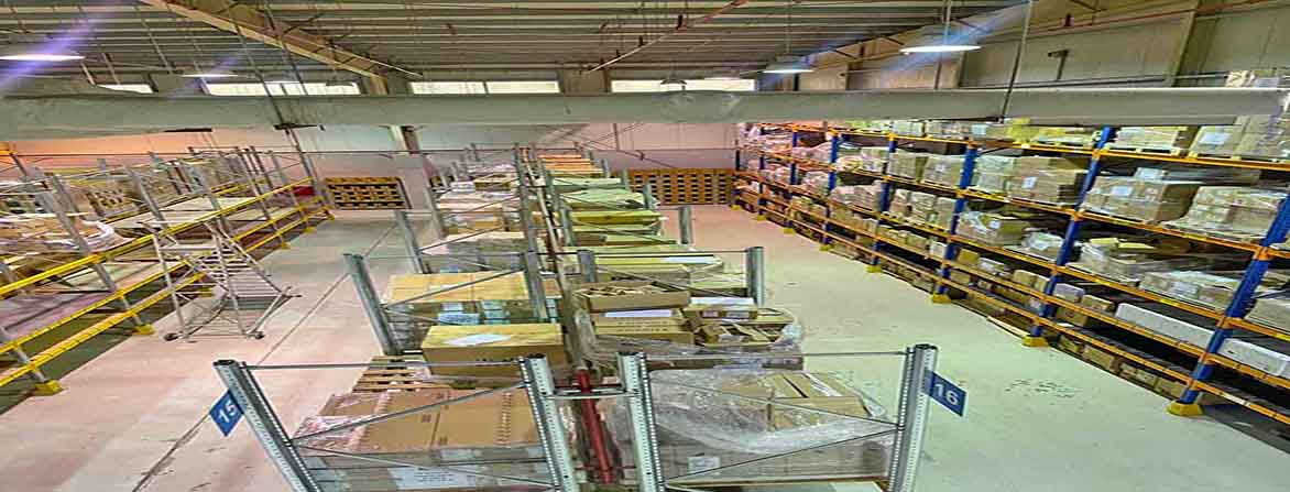 An overhead interior view of a vast industrial warehouse, showcasing its impressive 11-meter height. The concrete floor is clean, with long aisles separating rows of tall, heavy-duty metal racking systems filled with palletized goods wrapped in plastic. Bright overhead lighting illuminates the space, and exposed ductwork runs along the ceiling.