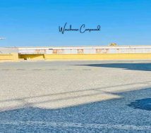 An outdoor shot of a large, empty asphalt paved warehouse compound or parking area under a clear blue sky. A yellow barrier wall runs along the back of the compound, and a construction crane is visible in the distance on the right. Long shadows stretch across the pavement, and a light pole stands on the left. The words "Warehouse Compound" are overlaid in the center of the sky.