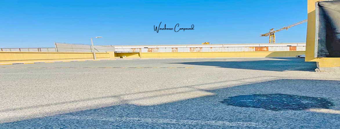 An outdoor shot of a large, empty asphalt paved warehouse compound or parking area under a clear blue sky. A yellow barrier wall runs along the back of the compound, and a construction crane is visible in the distance on the right. Long shadows stretch across the pavement, and a light pole stands on the left. The words "Warehouse Compound" are overlaid in the center of the sky.