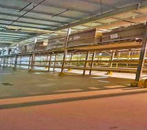 An expansive interior view of a well-lit warehouse with a high ceiling, polished concrete floor, and rows of heavy-duty metal shelving on both sides. Some shelves hold palletized goods. The far end of the warehouse appears to extend into the distance, and the "Samsproperties.com" logo is visible on the floor in the foreground.