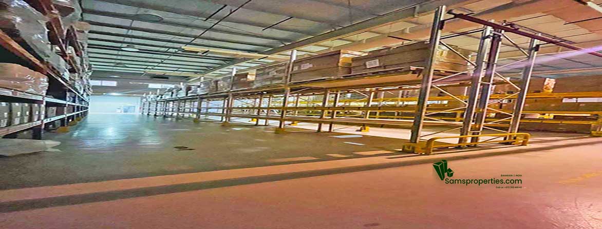 An expansive interior view of a well-lit warehouse with a high ceiling, polished concrete floor, and rows of heavy-duty metal shelving on both sides. Some shelves hold palletized goods. The far end of the warehouse appears to extend into the distance, and the "Samsproperties.com" logo is visible on the floor in the foreground.