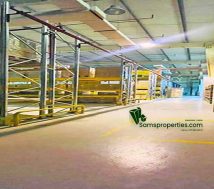 A wide-angle interior view of a modern, well-lit warehouse with a high ceiling, concrete floor, and extensive metal racking on both sides of a central aisle. Yellow safety markings are visible on the floor, and a red fire sprinkler pipe runs overhead. In the distance, more warehouse space can be seen. A "Samsproperties.com" logo is watermarked on the floor.