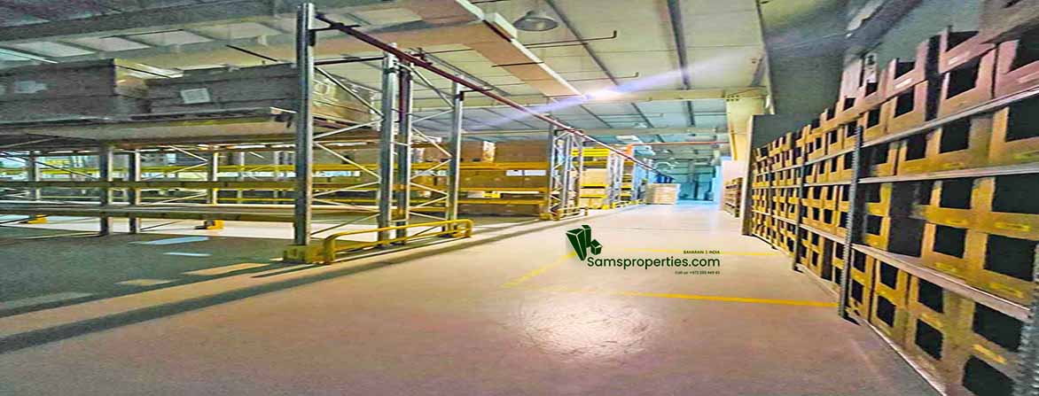 A wide-angle interior view of a modern, well-lit warehouse with a high ceiling, concrete floor, and extensive metal racking on both sides of a central aisle. Yellow safety markings are visible on the floor, and a red fire sprinkler pipe runs overhead. In the distance, more warehouse space can be seen. A "Samsproperties.com" logo is watermarked on the floor.