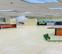 A spacious, well-lit office area within a warehouse, featuring a tiled floor, dropped ceiling with fluorescent lighting, several empty wooden desks with office chairs, filing cabinets, and a water cooler. Partitions separate different sections of the office, and a fire extinguisher is visible. The words "Office Space" are overlaid in the bottom left, and a "Samsproperties.com" logo with contact information is in the bottom right.