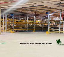 Inside a spacious, Civil Defence-approved warehouse for rent in Ras Zuwayed, Bahrain, featuring extensive racking, an 11-meter ceiling, high power load, multiple loading bays, and visible fire protection systems. A Samsproperties.com logo is present. A large warehouse of 2060 square meters (27986 Sqft). Warehouse for storage, Industrial workshop and factory.