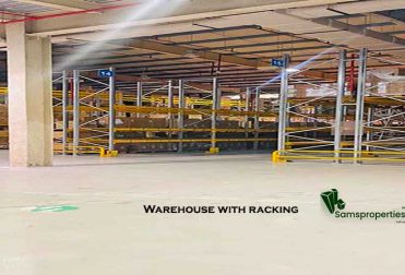 Inside a spacious, Civil Defence-approved warehouse for rent in Ras Zuwayed, Bahrain, featuring extensive racking, an 11-meter ceiling, high power load, multiple loading bays, and visible fire protection systems. A Samsproperties.com logo is present. A large warehouse of 2060 square meters (27986 Sqft). Warehouse for storage, Industrial workshop and factory.