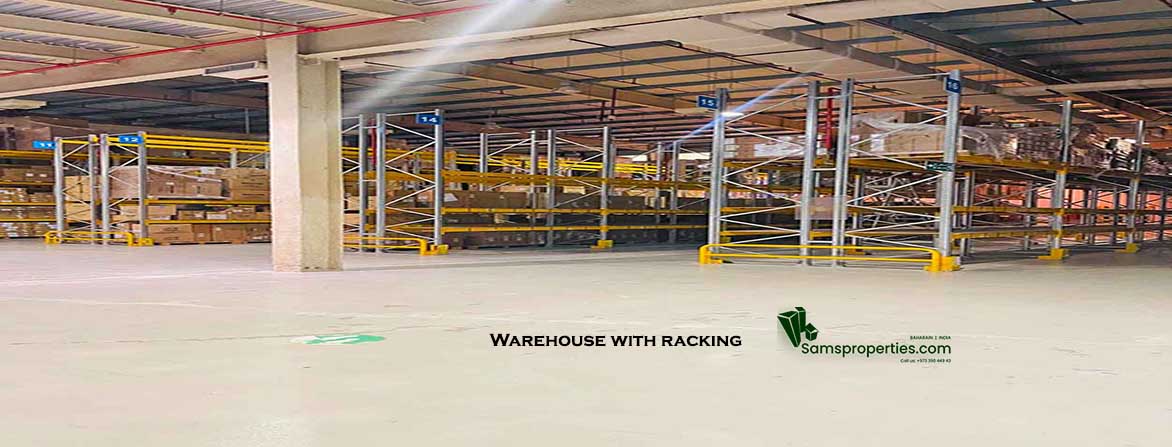 Inside a spacious, Civil Defence-approved warehouse for rent in Ras Zuwayed, Bahrain, featuring extensive racking, an 11-meter ceiling, high power load, multiple loading bays, and visible fire protection systems. A Samsproperties.com logo is present. A large warehouse of 2060 square meters (27986 Sqft). Warehouse for storage, Industrial workshop and factory.