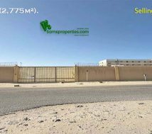 An image of a land for sale at Ras Zuwayed in Bahrain with a long, tan-colored boundary wall and a large metal gate in the centre. The wall is topped with barbed wire. In the background is a modern, light-colored building, and a paved road with a curb is in the foreground. The land is for sale to build a warehouse or workshop or factory.