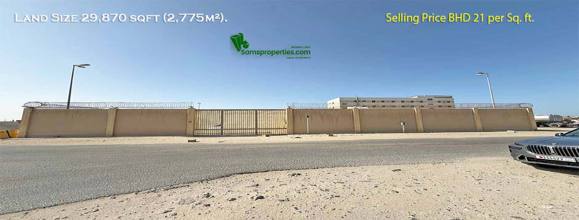 An image of a land for sale at Ras Zuwayed in Bahrain with a long, tan-colored boundary wall and a large metal gate in the centre. The wall is topped with barbed wire. In the background is a modern, light-colored building, and a paved road with a curb is in the foreground. The land is for sale to build a warehouse or workshop or factory.
