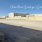 A wide-angle photo of a large, empty industrial land for sale at Ras Zuwayed in Bahrain under a clear blue sky. In the foreground is a paved area with a curb and sidewalk, while a beige warehouse with two large garage doors is visible in the middle ground. A taller, multi-story building is in the background. The lot is enclosed by a high wall topped with barbed wire on both the left and right sides. This land is proposed to build an industrial shed for storage or a heavy workshop or a factory.