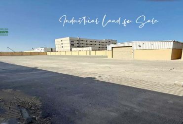 A wide-angle photo of a large, empty industrial land for sale at Ras Zuwayed in Bahrain under a clear blue sky. In the foreground is a paved area with a curb and sidewalk, while a beige warehouse with two large garage doors is visible in the middle ground. A taller, multi-story building is in the background. The lot is enclosed by a high wall topped with barbed wire on both the left and right sides. This land is proposed to build an industrial shed for storage or a heavy workshop or a factory.