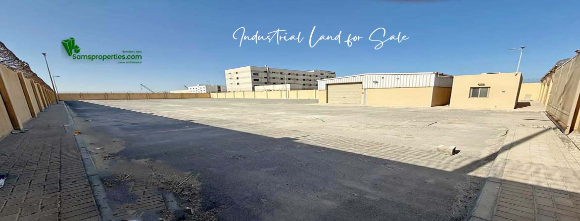 A wide-angle photo of a large, empty industrial land for sale at Ras Zuwayed in Bahrain under a clear blue sky. In the foreground is a paved area with a curb and sidewalk, while a beige warehouse with two large garage doors is visible in the middle ground. A taller, multi-story building is in the background. The lot is enclosed by a high wall topped with barbed wire on both the left and right sides. This land is proposed to build an industrial shed for storage or a heavy workshop or a factory.