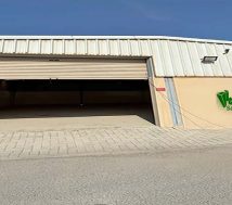 A photograph of an empty warehouse on an industrial land in Bahrain with a large, partially open rolling shutter door. The warehouse walls are beige, and the roof is a white corrugated metal. The ground in front of the warehouse is paved with light-colored bricks.