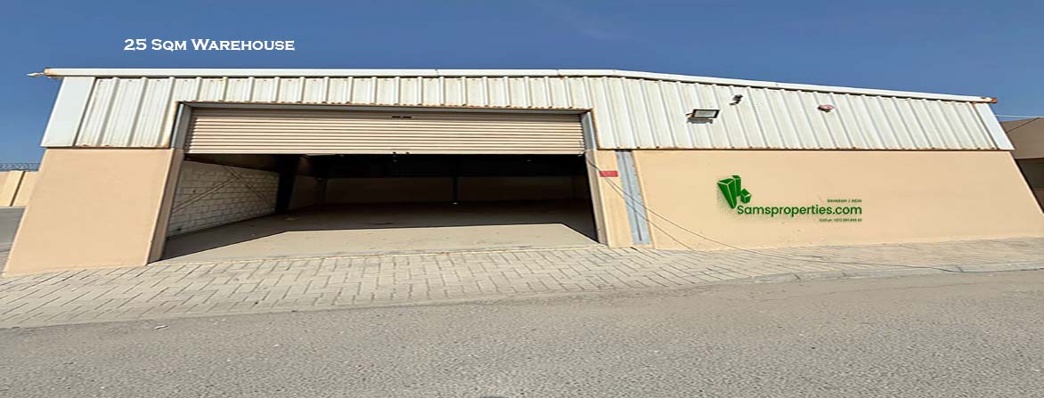 A photograph of an empty warehouse on an industrial land in Bahrain with a large, partially open rolling shutter door. The warehouse walls are beige, and the roof is a white corrugated metal. The ground in front of the warehouse is paved with light-colored bricks.