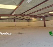 The interior of an empty warehouse shows a concrete floor, a high ceiling with exposed metal beams, and corrugated metal walls. Skylights in the roof and fluorescent lights provide illumination.